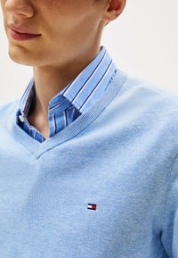 Zbliżenie szyi i twarzy osoby noszącej jasnoniebieski sweter w serek na niebieskiej i białej w paski koszuli z kołnierzykiem. Widoczny mały logo.