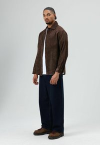 Bruine corduroy overhemdjas met knoopsluiting, gecombineerd met een wit T-shirt en donkerblauwe wijde broekspijpen. Bruine veterlaarzen met dikke zolen.