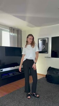 White t-shirt, black pinstriped trousers, and black sandals; hair down, featuring hoop earrings and a watch; indoors with a TV in the background.