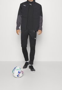 Chaqueta deportiva negra con parte superior acolchada, mangas grises, combinada con pantalones negros y zapatos de fútbol. Hay una pelota de fútbol multicolor cerca.