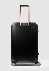 Schwarzer Hartschalenkoffer mit vertikal geriffeltem Design, ausziehbarem Griff in Roségold und vier Rädern an der Basis für Mobilität.