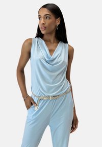 Top sin mangas de color azul claro con un escote drapeado, fabricado en un tejido suave, complementado con un cinturón trenzado blanco. La modelo posó con una mano en el bolsillo.