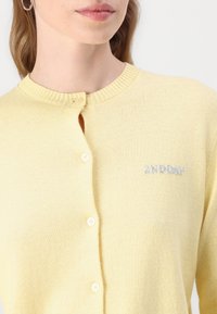 Light yellow cardigan with a round neckline, featuring a button closure and subtle embroidered branding. Soft texture and simple design.