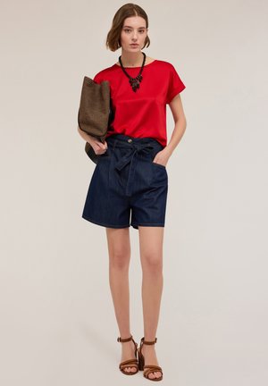 Rode satijnen top met korte mouwen gecombineerd met donkerdenim hoge taille shorts. Model houdt een gestructureerde bruine tas vast. Bruine sandalen met banden maken de look compleet.
