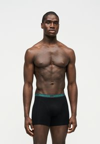 Μυώδης άνδρας με σκούρο δέρμα, όρθιος, φορώντας μαύρα ανδρικά εσώρουχα Björn Borg με πράσινη μέση, μπροστά σε απλό γκρι φόντο.