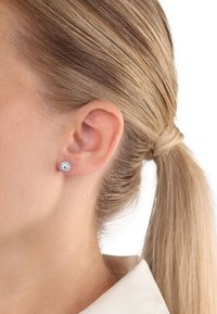 Pendientes de botón plateados en forma redonda con un diseño floral realzado con múltiples piedras preciosas transparentes, que complementan un peinado elegante y una blusa en tonos neutros.