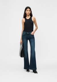 Γυναίκα που φοράει μαύρο αμάνικο τοπ και σκούρο μπλε παντελόνι καμπάνα, κρατώντας μαύρη τσάντα ώμου, στέκεται μπροστά σε λευκό απλό φόντο.