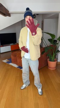 Beige jakke, hvit skjorte, grå bukser og sølvfargede joggesko. Burgunderfargede hansker og en blå hatt med dusk. Innendørs miljø med planter.