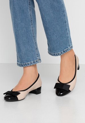 Beige og sorte lavhælede sko med sorte sløjfer, båret med blå jeans, vist fra midt på læggen til gulvet på en lys grå overflade.
