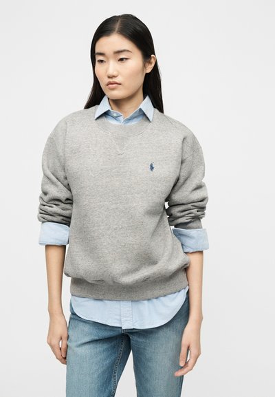 Νεαρή γυναίκα με μακριά σκούρα μαλλιά, φορώντας γκρι φούτερ πάνω από ανοιχτόγαντζο μπλουζάκι και μπλε τζιν, στέκεται μπροστά σε απλό φόντο.