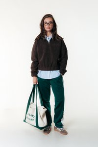 Pull en molleton marron à demi-zip, chemise rayée bleue en dessous, pantalon de sport vert et chaussures marron. Tient un sac fourre-tout crème avec des poignées vertes.