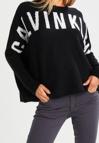 Svart oversized tröja i mjuk trikå med stor vit text "CALVIN KLEIN" på framsidan. Ribbad kant vid ärmar och nederkant.