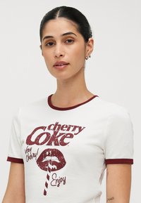 Bela bombažna majica s temno rdečimi rebrastimi ovratnikom in rokavi. Ponuja grafični tisk z logotipom "cherry Coke", rdečimi ustnicami in slamo.