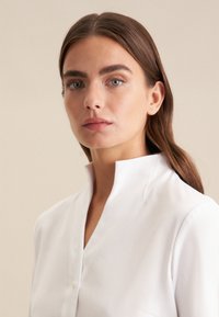 Wit overhemd met een staande kraag, met een gladde textuur en minimale knoopdetails. Zachte verlichting accentueert de stof.
