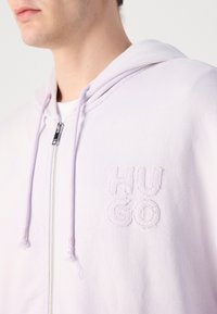 Hanorac cu fermoar de culoare mov deschis, din bumbac, având un glugă cu șnur și un logo brodat "HUGO" pe partea stângă a pieptului. Textură netedă.