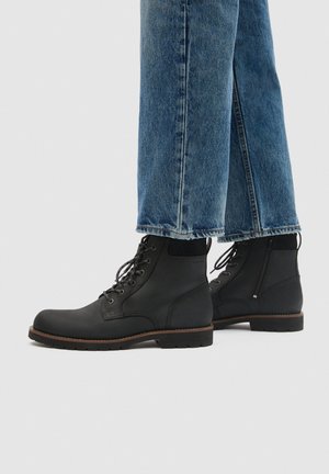 Bottines noires à lacets et à semelles épaisses, portées sous un jean bleu à jambes larges.