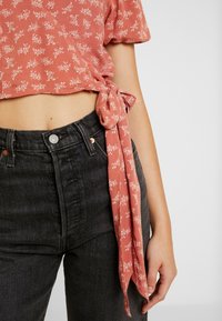Femme portant un jean noir taille haute et un haut court à manches courtes, rose à motifs floraux, noué à la taille.