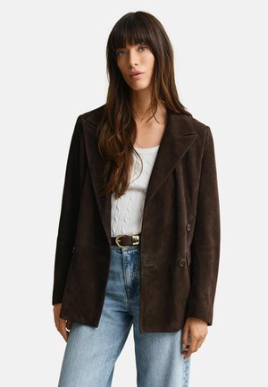 Frau mit langen braunen Haaren, die einen dunkelbraunen Wildleder-Blazer, einen weißen Strickpullover mit Zopfmuster, hellblaue Jeans und einen schwarzen Gürtel mit goldener Schnalle trägt.