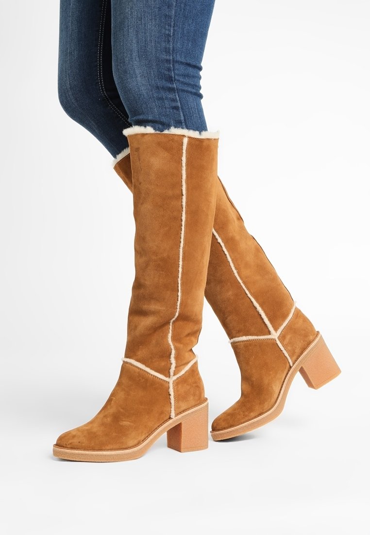 Personne portant de hautes bottes en suède marron clair avec des surpiqûres blanches et une doublure en fourrure, associées à un jean skinny bleu sur fond blanc.