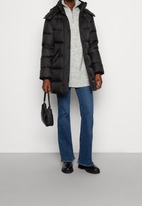 Zwarte pufferjack met capuchon, ritsluiting en knopenaccenten. Gecombineerd met een grijs gebreid topje, blauwe flared jeans en zwarte schoenen.