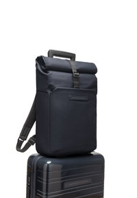 Sac à dos roll-top bleu marine foncé en matériau imperméable avec des sangles ajustables et une poche avant, positionné sur une valise brillante.