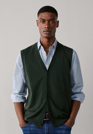 Jonge man met kort haar draagt een donkergroene buttonvest over een lichtblauw shirt met opgerolde mouwen, staand met handen in de zakken.
