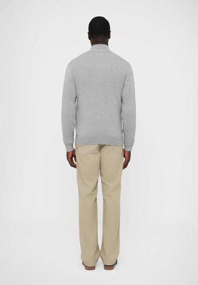 Szürke bordázott kötött pulóver magas gallérral, bézs nadrággal párosítva, hátulnézetben. Puha textúra és testhezálló szabás.