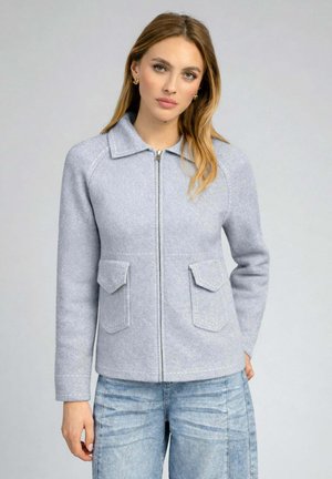 Femme portant une veste zippée gris clair avec col et poches à rabat, associée à un jean bleu clair, debout devant un fond uni.