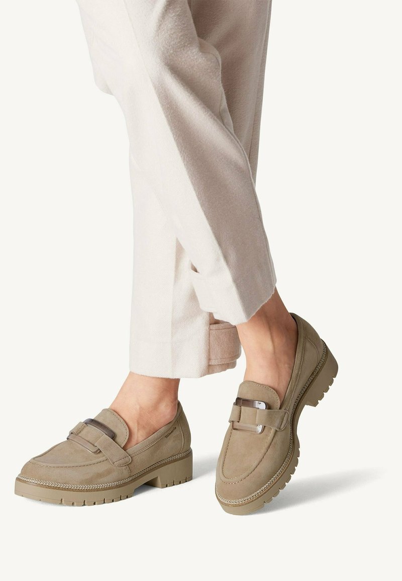 Mocassins en daim beige avec semelles épaisses et détail de boucle métallique, portés avec un pantalon court de couleur claire.