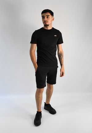 Camiseta negra ajustada con un pequeño logo, combinada con pantalones cortos negros y zapatillas deportivas negras. Textura suave, diseño casual.
