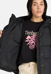 Zwarte pufferjack met ritssluiting en grote zakken, gedragen over een zwart graphic T-shirt met een roze bloemendessin.