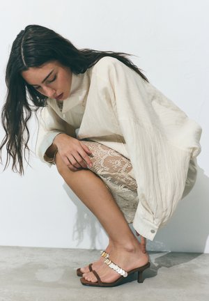 Žena s dlhými tmavými vlasmi, ktorá nosí krémovú voľnú bundu, čipkovanú sukňu a otvorené sandále na podpätku, kľačí s rukou na kolene.