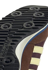 Brun mocka och vinröd textil sneakersula med svart gummiträd, inklusive cirkulära greppmönster och en vit accent på sidan.