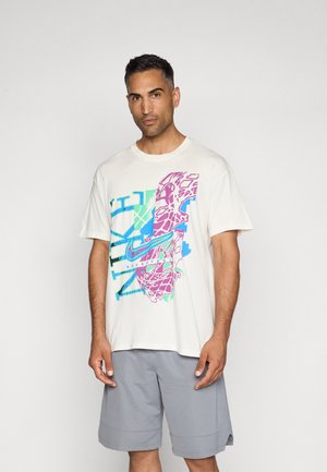 Tricou de culoare crem cu un grafic mare și colorat în roz, albastru și verde, purtat cu pantaloni scurți gri, având o croială lejeră și mâneci scurte.
