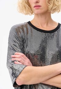 Haut en sequins argentés avec une coupe ample, des manches courtes et un col côtelé noir. La texture est brillante et réfléchissante, mettant en valeur un motif texturé.