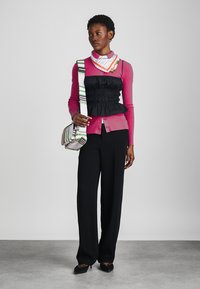 Sort rynket top lagd over en pink tætsiddende cardigan, båret med brede sorte bukser. Akkentueret med en stribet skuldertaske og tørklæde.