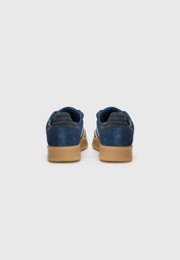 Zapatillas de ante azul con una suela de goma, adornadas con acentos en azul marino oscuro y tres rayas blancas a los lados. Talón texturizado y cuello acolchado.