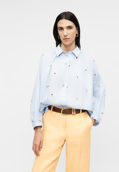 Femme portant une chemise ample à rayures bleu clair avec de petits imprimés floraux, un pantalon beige taille haute, une ceinture marron, des boucles d'oreilles créoles en or.
