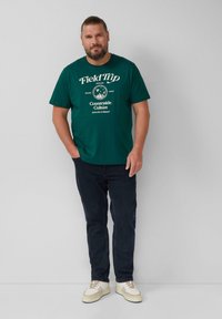Man met een baard en teruggebonden haar, gekleed in een donkergroene T-shirt met de tekst "Field Trip Countryside Culture", donkere spijkerbroeken en witte sneakers, staande tegen een grijze muur.
