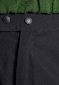 Gros plan sur un pantalon en tissu noir avec deux boutons noirs sur la ceinture et un vêtement en maille verte par-dessus.