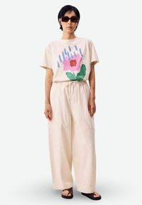 Mujer con pantalones beige holgados, camiseta crema con estampado floral en rosa y verde, gafas de sol grandes y sandalias negras, de pie frente a un fondo blanco.