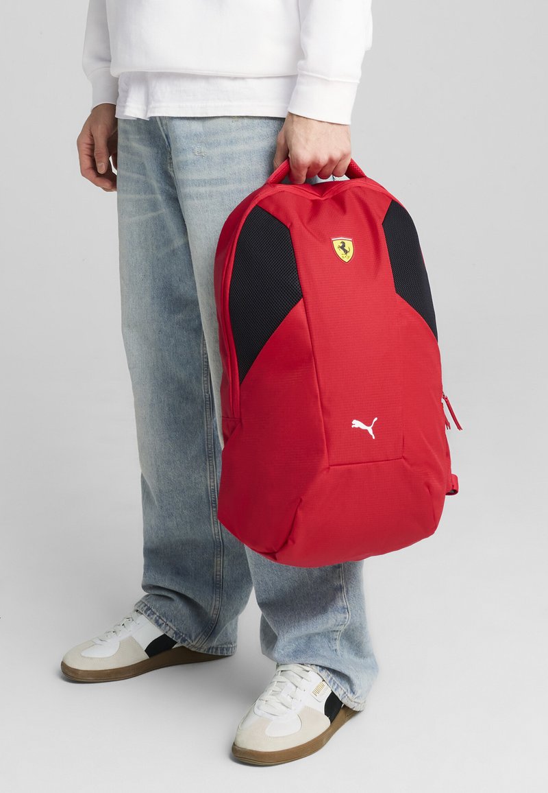 Zaino rosso con dettagli in rete nera, logo Puma ed emblema Ferrari. Caratterizzato da un scomparto con cerniera e una robusta maniglia per il trasporto.