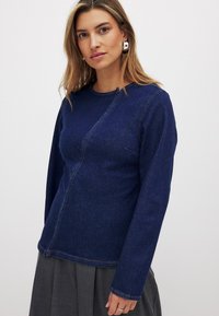 Tumši zila džinsa garā piedurkne augšdaļa ar apaļu kaklu, kurai ir izliekti šuvi un viegla tekstūra. Šuvju akcenti gar malām.