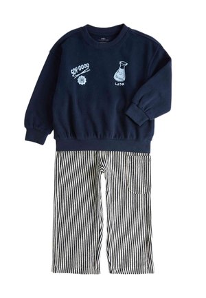 Junger Junge trägt einen marineblauen Pullover mit weißen Grafiken und schwarz-weiß vertikal gestreifte Hosen, steht mit auseinander stehenden Beinen vor einem einfarbigen Hintergrund.