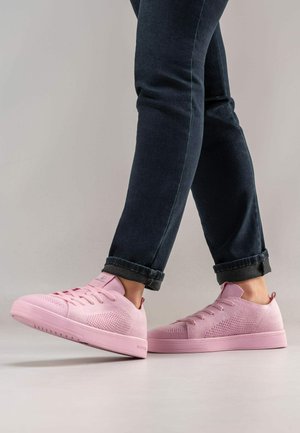Persoon die donkere omgeslagen jeans en lichtroze gebreide sneakers draagt, met één voet licht van een gladde grijze vloer opgetild.