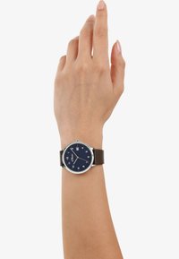 Bruin leren strap horloge met een ronde zilveren kast, blauwe wijzerplaat, witte cijfers en een datumnaald op 3 uur. Hand omhoog, horloge is zichtbaar.