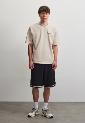 Junger Mann steht vor einer schlichten Wand, trägt ein beige oversized T-Shirt, schwarze Sportshorts, weiße Socken und weiße Sneakers.