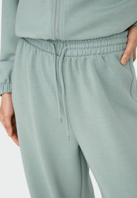 Nahaufnahme von hellgrünen, elastischen Hose mit Gummizug und Schnürung, mit einem Reißverschluss oben und einer Hand, die in der Tasche ruht.