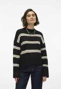 Maglione nero lavorato a maglia con strisce orizzontali bianche, caratterizzato da un colletto alto e maniche larghe, abbinato a jeans scuri. Tessuto texturizzato visibile.