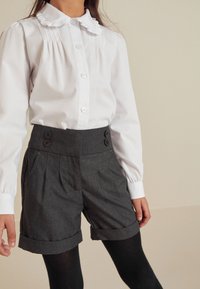 Witte langemouwen shirt met een ruches-kraag, gecombineerd met hoge grijze shorts met knopen en opgerolde zoom. Zwarte panty’s maken de outfit af.